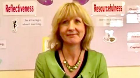 Christine Baker is seen in front of a school noticeboard showing a display about learning strategies. She has blonde hair with a fringe and is wearing a green jacket with a black top and a necklace of green beads.