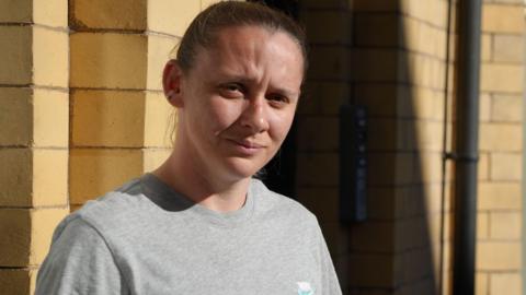 Elissa Hubbard wearing a grey Nike T-shirt and looking at the camera. She is standing in front of her home which is yellow brick and a window is visible in the background