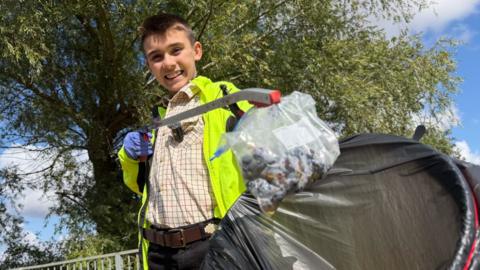 Young boy with brown hair, wearing a smart plaid shirt and yellow hi-vis smiles to camera holding a bag and litter picker.
