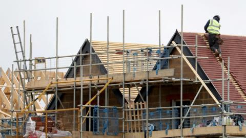 A generic shot of housebuilding, with a man tiling a roof and scaffolding against a partially built house.