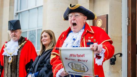 An older male town crier cries while reading from an embroidered scroll. He is wearing a traditional town crier hat and outfit as he stands in front of the entrance to a town hall. There two other ceremonial figures - a man and a woman - stood beside him.