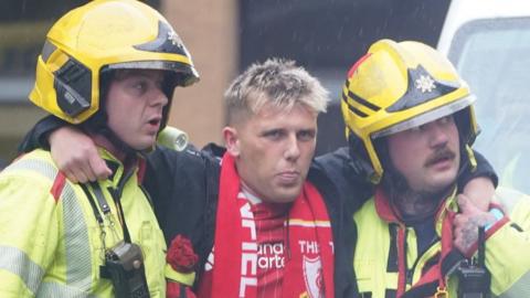 A young man with bleached blonde hair is wearing a Liverpool FC top and scarf is being held up by two firemen who are wearing yellow helmets and jackets.