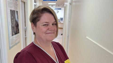 Helen Clyde is standing side-on and smiling at the camera. She has short, blonde hair and is wearing a red and white uniform, with a yellow name tag. The hospital corridor walls behind her are white and and have posters with medical information on them.