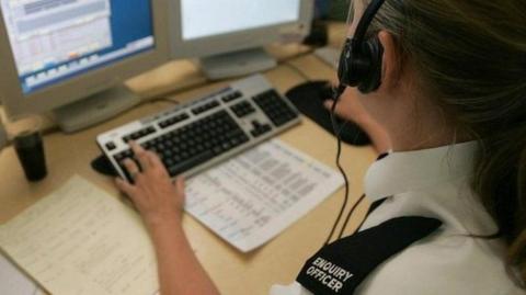 A police call handler sits at a desk working on a computer with several screens in front of her as well as a keyboard and mouse. She wears a headset with a microphone, white shirt with a black lapel on the shoulder with the words enquiry officer on it.