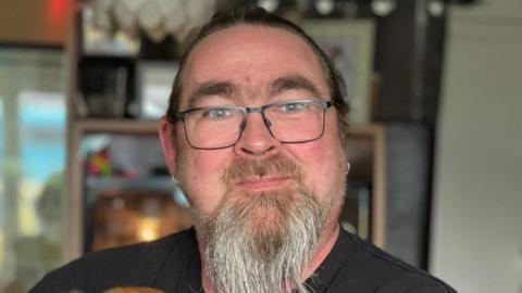 Allan Batcock with brown hair and brown and grey beard wears black glasses, black t-shirt and silver earrings. He is in his house with a bookcase behind him and patio doors leading to the garden blurred out. He is slightly smiling.