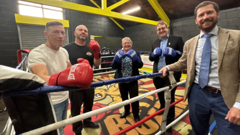 Three men and a woman standing in a boxing ring, and one man standing just outside it. The ones inside are wearing boxing gloves and they are all smiling.