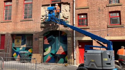 Two men on a cherry picker removing the bas-relief carving from the outside of the red brick Victorian building. Below them are two doorways covered in wooden panels painted with abstract artwork.