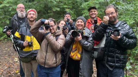 A group of adults huddle together. They are soaked through in waterproof clothes and holding up cameras.