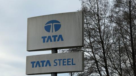 A close-up of a sign for Tata Steel. It is a white board with Tata Steel written in blue, with trees behind it.