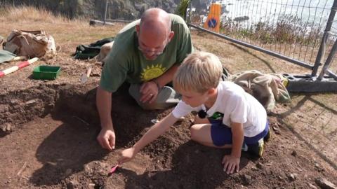 An archaeological excavation in progress, featuring two individuals — an adult and a child — kneeling on the ground and carefully digging with small tools. The adult is wearing a green shirt with a yellow sun design, and the child is dressed in a white shirt and blue shorts. 