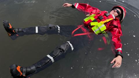 A man dressed in a red waterproof jacket, red helmet and black trousers and black and orange boots floating on murky-looking water.