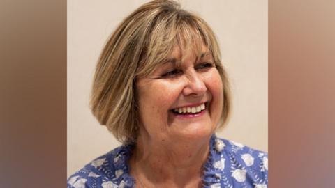 Janine Roebuck, who has a light brown blonde streaked bob with a fringe. She is smiling broadly, turning her head to the right and is wearing a blue and white cotton top 