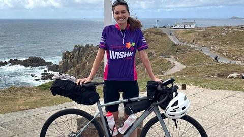 A woman with long brown hair tied back and black cycle glasses on her head wearing a purple top that says 'Wishh' and black cycle shorts. She is standing in front of a bike and holding its saddle and handlebars which have a helmet and water bottles on it. Behind her is a cliff with the sea at the bottom.