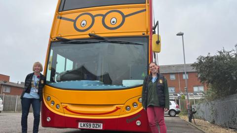 Mandy Potter and Lindsey Beddow stand either side of a double-decker bus which is painted with a yellow front with eyes and glasses above the windscreen.