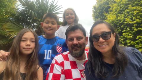 Robin, 14, Max, 12, Zoe, 10, and their parents Mladen and Jaime, are outside in a garden, with Max wearing a blue football shirt, and his father wearing a red and white football shirt.