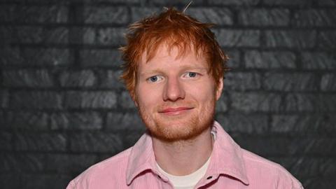 Ed Sheeran wearing a pink jacket and white top. 
