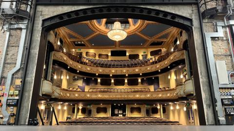 Inside Jersey Opera House. The photographer is stood on the venue's stage looking out towards the auditorium, which has gold and blue seating and walls. A large chandelier is hanging above the room. 