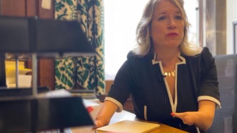 A woman with blonde hair and wearing a dark dress is sat in an office chair in front of a desk. She has foreshortened arms with only one digit on each hand. A notepad is on the desk in front of her and a stack of office letter trays. Behind her there is a window with light coming into the room and a white and green patterned curtain.