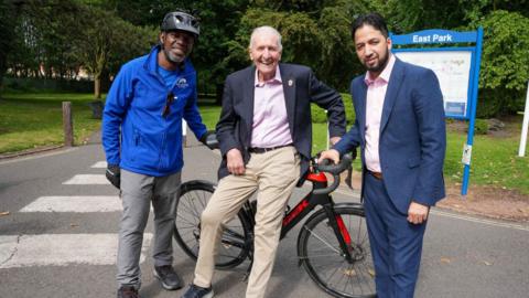 Three men standing in front of a racing bike on a path next to a park. A sign in the background reads East Park. A man at the centre with white hair in beige trousers, pink shirt and blazer leans against it, holding the handlebar. A man on his left is in a blue jacket and cycling helmet, and on his right a man in a navy suit with pink shirt. 