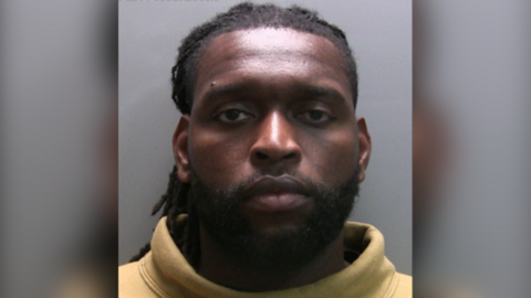 Custody photo of Emmanuel-Thomas. He looks serious and is wearing a yellow turtle neck jumper.