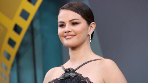 Actress Selena Gomez smiles at premiere in Los Angeles. She wears a black dress with exposed shoulders. Her long dark hair is held back with a hairband.