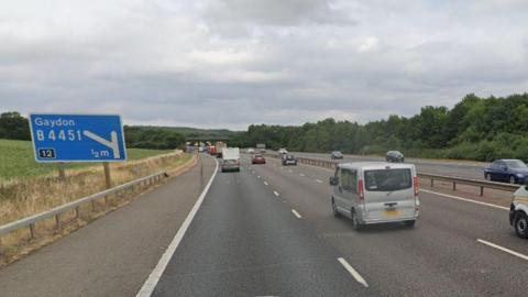 A Google Street View image of the M40 in Warwickshire. The northbound carriageway road sign for Gaydon junction 12 can be seen and there are cars, lorries and vans driving on both sides of the route.