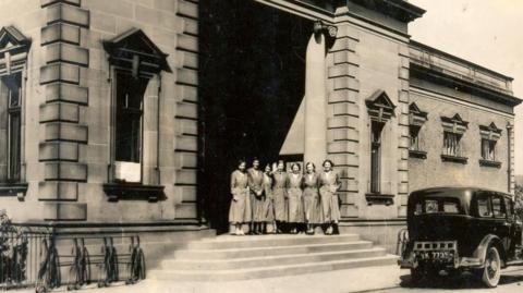 A black and white photo showing seven women standing outside Gateshead Central Library. The library is large and made of stone.