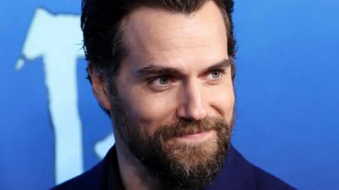 A man with grey eyes, a strong jaw and brown beard, smiles in front of a blue background.