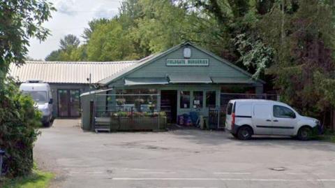A single-storey grey building which has Fieldgate Nurseries written above its door. In front of the doors and windows are plants and flowers. To the right of the building is a while van. In front of it is a carpark. 