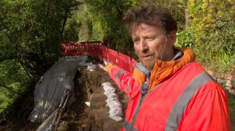 Tristen Dodd pictured on the Railway Walk. The landslip is visible behind him. He is gesturing towards it. He's wearing an orange hi-vis jacket. He has short brown hair and a beard. 