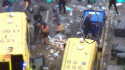 A still from police helicopter footage shows several men, including one highlighted in an oval, in a street full of debris caused by a riot. There are three men gathered around one of the two police vans in shot and several domestic bins and one industrial bin overturned in the street