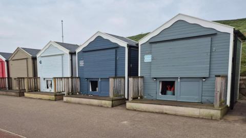 Four coloured beach huts in a row painted light brown, pale blue, dark blue and dark green. Each has a small decking to its front. Each has a panel missing, which is how the intruder gained access to the space. 