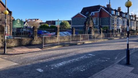 A faded zebra crossing goes across a worn road. It has Belisha beacons on each side of it. There is a green sign denoting the Old School Surgery while a railing on the far side of the road stretches diagonally into the background towards a row of terraced houses.