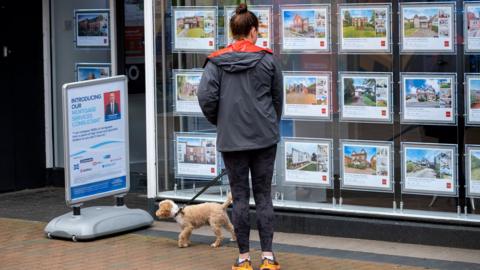 A woman looks in teh window of an estate agent . She is wearing sporty clothes and has a small dog on a lead with her