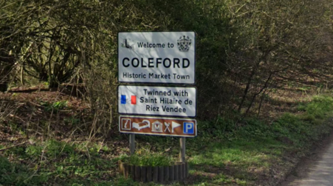 A road sign that says 'Welcome to Coleford, Historic Market Town'. It is on the side of a road which is out of the picture. Behind the sign is a number of trees and bushes. 