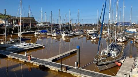 More than a dozen small boats moored at Whitehaven Harbour. The water is a dark orange/brown colour. The boats are moored along wooden boardwalks. The sky beyond is a deep blue.