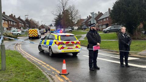 Two police officers stood behind a cordon in a road. A police car is behind them and houses line the street