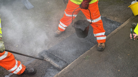 A close up view of workmen in bright orange overalls holding a bucket of tar and a tool to fix a pothole on the road