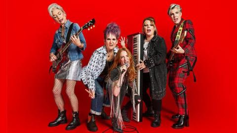 The five main cast members of Riot Women pose against a red background. On the left of the image Amelia Bullmore wears a denim jacket and silver skirt while playing a red guitar. Lorraine Ashbourne holds a set of drumsticks as she crouches next to Rosalie Craig, who is holding a microphone. Joanna Scanlan wears a long, black denim jacket and black trousers. She is holding a keyboard. On the right of the picture Tamsin Greig is wearing a red tartan jacket and matching trousers. She is holding a bass guitar.
