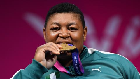 A headshot shows Folashade Oluwafemiayo of Team Nigeria posing for a photo during the medal ceremony for the women's heavyweight para-powerlifting at the Commonwealth Games by biting on her gold medal which is on a pink and purple ribbon around her neck. She wears a green tracksuit top with white trim and blurred in the background is a pink hoarding showing part of the Glasgow 2026 logo in white