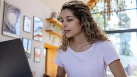 A stock image of a woman sat at a laptop. She has brown curly hair with some blond, and is wearing a purple t-shirt. 