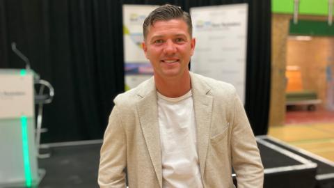 Luke wears a relaxed stone coloured jacket over a white t-shirt and is smiling for the camera. A black stage is in the background.