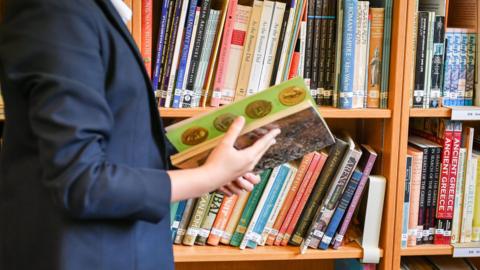 Someone wearing a blazer, they carry an old book and look at it, and there are shelves of history books in the background.
