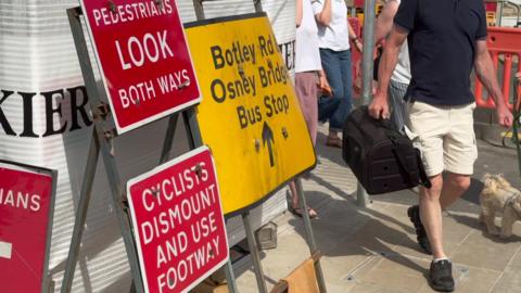 People walking near sings on Botley Road  signs reading "Pedestrians look both ways", "Cyclists dismount and use footway" and "Botley Road Osney Bridge Bus Stop".