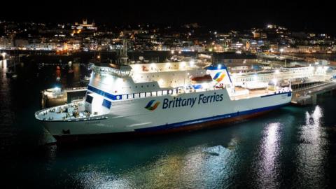 A large white and blue Brittany Ferries ship is docked in a harbour at night, brightly lit against dark water. City lights from buildings on the hillside glow in the background, reflecting across the harbour, while the ferry’s ramps and terminal structures extend alongside the vessel.