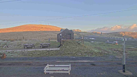 The Bungalow, you can see frost on the ground, the peak of a hill is on the left, with a road on the right and a small building is in the foreground.