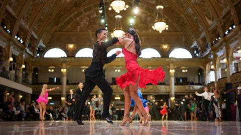 A ballroom dancing couple wearing black and red dancing in the centre of a large dancefloor.