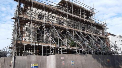 A three-storey building is covered in scaffolding. There is a wooden fence in front of it.