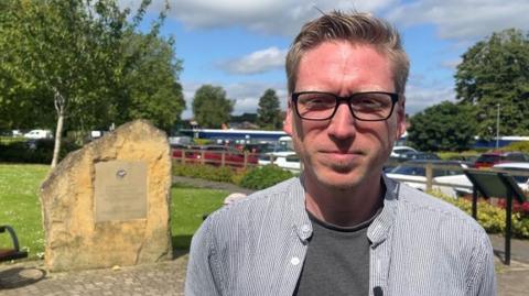 Dan Boatright-Greene is stood outside wearing an open necked shirt and a grey T-shirt underneath. He is also wearing glasses. Behind him on the left is a large honey-coloured stone with a plaque attached to it. In the background there is a tree and a car park, It is a sunny day.