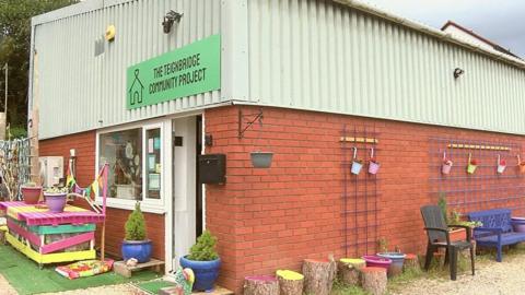A low-level industrial building with a sign in green and writing on it with a numbver of pot plants, chairs and other coloured garden paraphernalia outside.